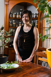 a white woman in her 30s stands in front of her apothecary shelf, filled with jars of herbs
