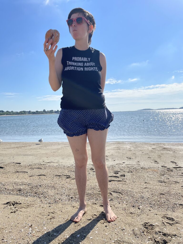 a white woman with short hair stands on a beach eating a donut. Her shirt reads "probably thinking about abortion rights"