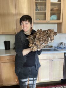 a white woman in her mid 30s holding up a large hen of the woods mushroom in her kitchen